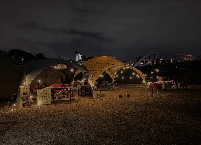 夜の河川敷に設営された、ライトアップされた大型テントと焚き火会場の全景